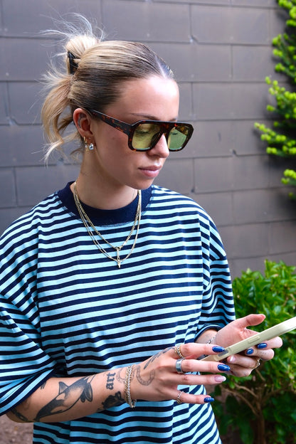 junge Frau mit Handy in der hand trägt Sonnenbrille mit braunem rahmen und großen grünen Gläsern die aber nur leicht verdunkelt sind