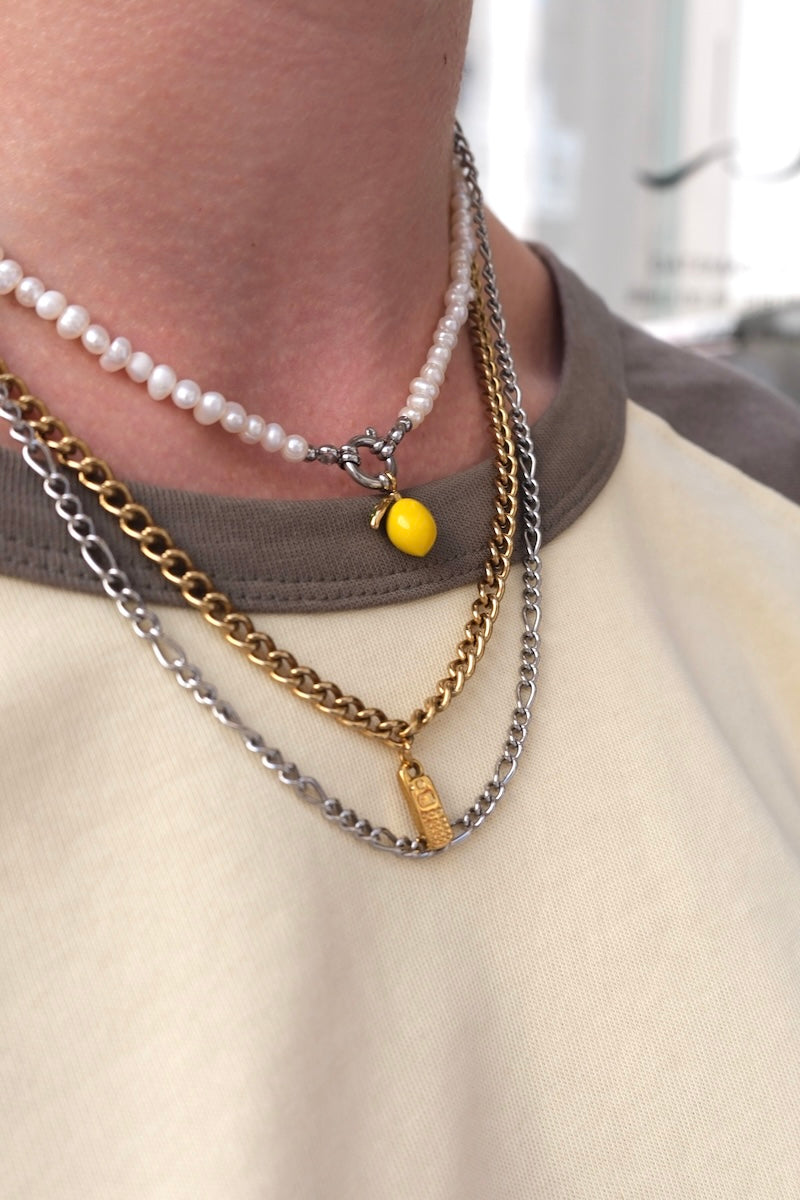 zu sehen ist der obere brust und Halsbereich einer person mit mehreren übereinander getragenen Halsketten, bei einer der ketten ist ein goldfarbiger charm in form eines mobiltelefon eingehängt und bei einer anderen kette ist ein charm in form einer Zitrone eingehängt