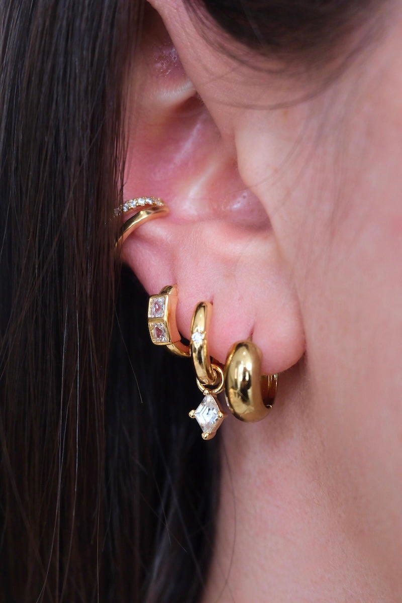 zu sehen ist ein ohr mit drei hoops und einem earcuff. im ersten loch ist ein dicker Basic hoop. im zweiten loch ein schmaler Basic hoop mit einem stein drauf und einem Diamant charm der eingehangen ist. im dritten loch sieht man ein dickeren hoop mit zwei dicken Diamant steinen drauf und oberhalb am ohr ist ein Doppel earcuff die untere Seite ist clean gold und die Ober Seite ist mit zirkonia steinen besetzt