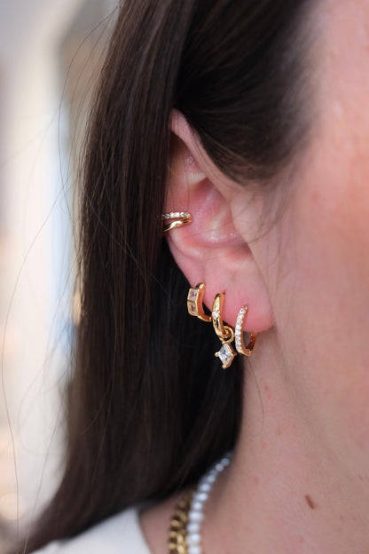 zu sehen ist ein ohr mit drei hoops und einem earcuff. im ersten loch ist ein dicker Basic hoop. im zweiten loch ein schmaler Basic hoop mit einem stein drauf und einem Diamant charm der eingehangen ist. im dritten loch sieht man ein dickeren hoop mit zwei dicken Diamant steinen drauf und oberhalb am ohr ist ein Doppel earcuff die untere Seite ist clean gold und die Ober Seite ist mit zirkonia steinen besetzt