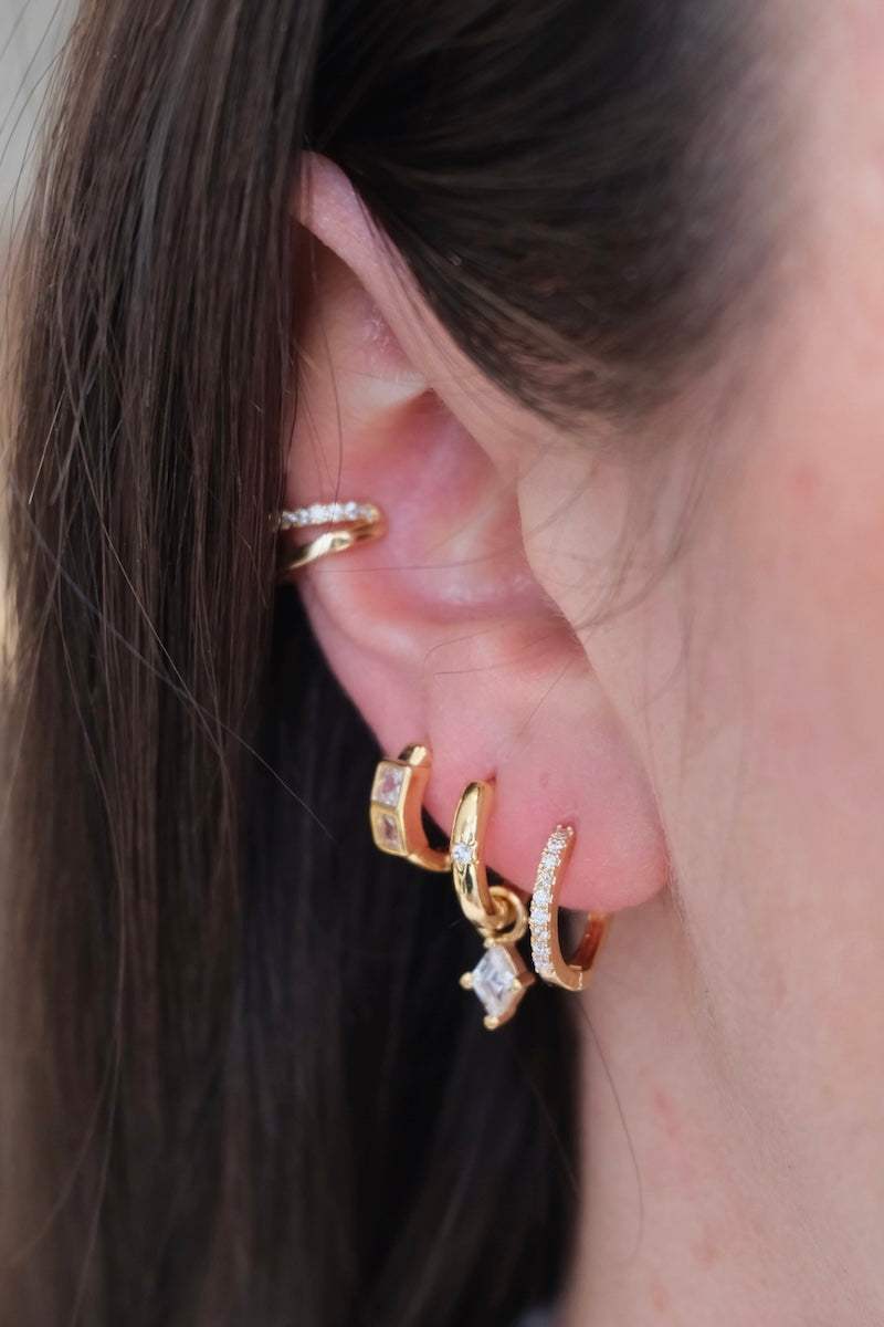 zu sehen ist ein ohr mit drei hoops und einem earcuff. im ersten loch ist ein dicker Basic hoop. im zweiten loch ein schmaler Basic hoop mit einem stein drauf und einem Diamant charm der eingehangen ist. im dritten loch sieht man ein dickeren hoop mit zwei dicken Diamant steinen drauf und oberhalb am ohr ist ein Doppel earcuff die untere Seite ist clean gold und die Ober Seite ist mit zirkonia steinen besetzt