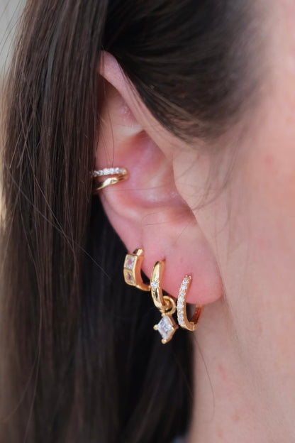 zu sehen ist ein ohr mit drei hoops und einem earcuff. im ersten loch ist ein dicker Basic hoop. im zweiten loch ein schmaler Basic hoop mit einem stein drauf und einem Diamant charm der eingehangen ist. im dritten loch sieht man ein dickeren hoop mit zwei dicken Diamant steinen drauf und oberhalb am ohr ist ein Doppel earcuff die untere Seite ist clean gold und die Ober Seite ist mit zirkonia steinen besetzt