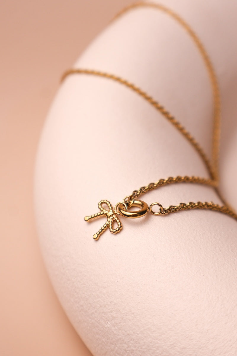 Filigrane goldene Zopfkette verbunden mit einem runden goldenen Ringcharm an dem eine Schleifencharm hängt
